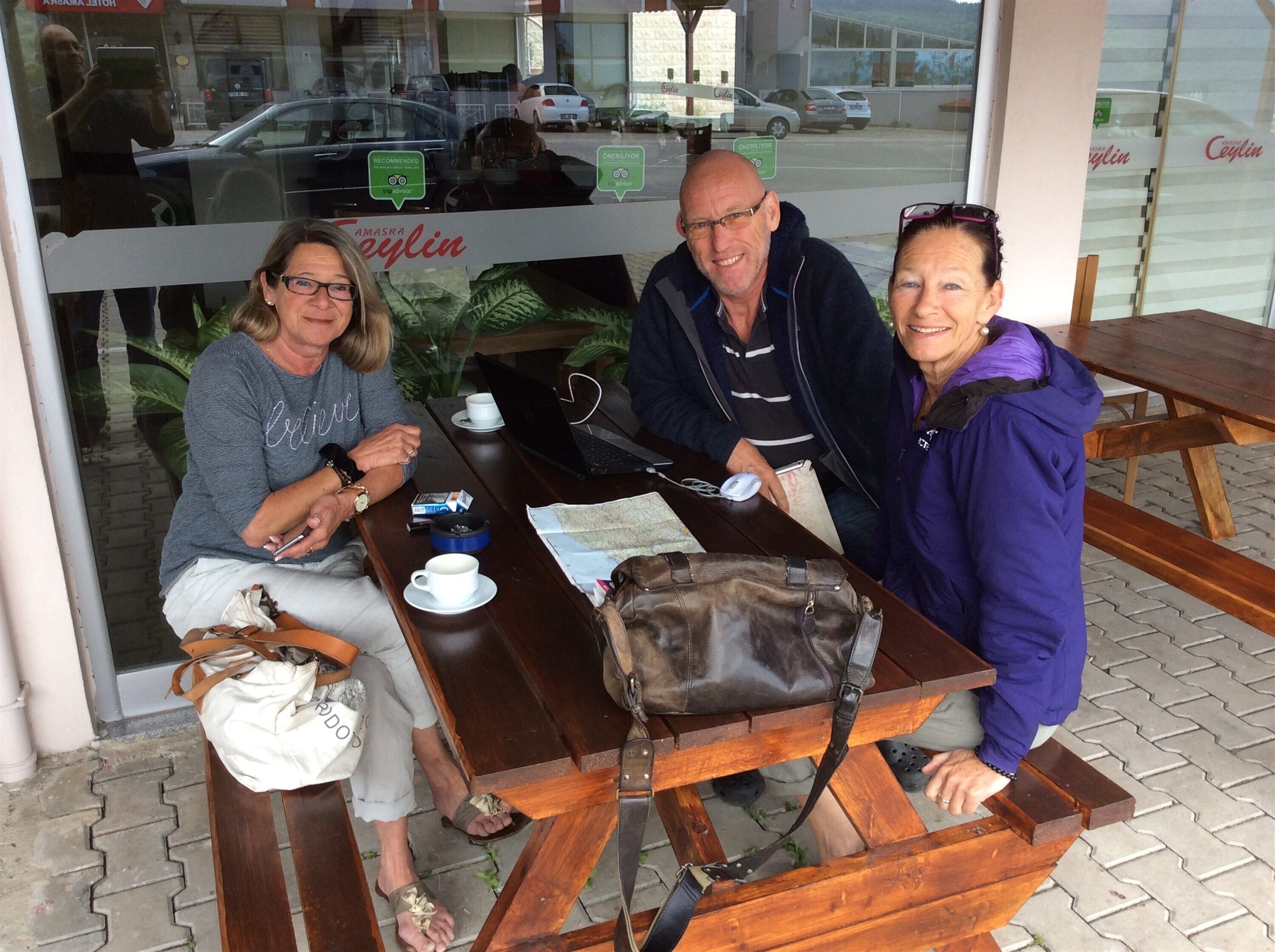 Three people seated at an outdoor wooden table with a laptop, a cup, and a handbag.