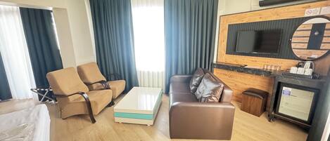 A hotel room with a sofa, a coffee table, a TV mounted on a wooden wall, and a small kitchenette.