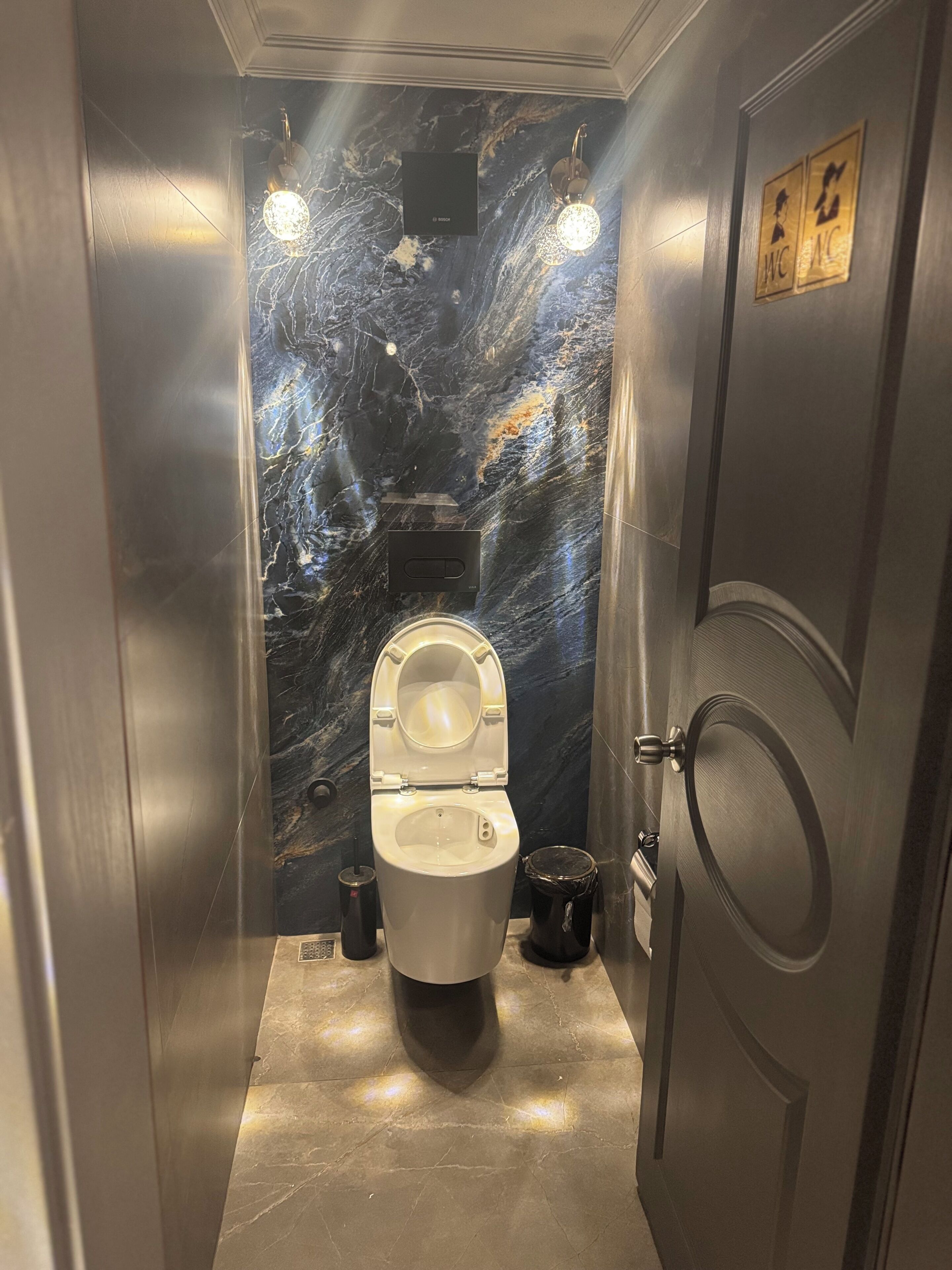 A compact, modern bathroom with a marble wall, a toilet, and a trash bin.