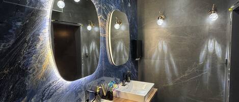 Un baño moderno con dos lavamanos, paredes de mármol y ducha empotrada con luces.