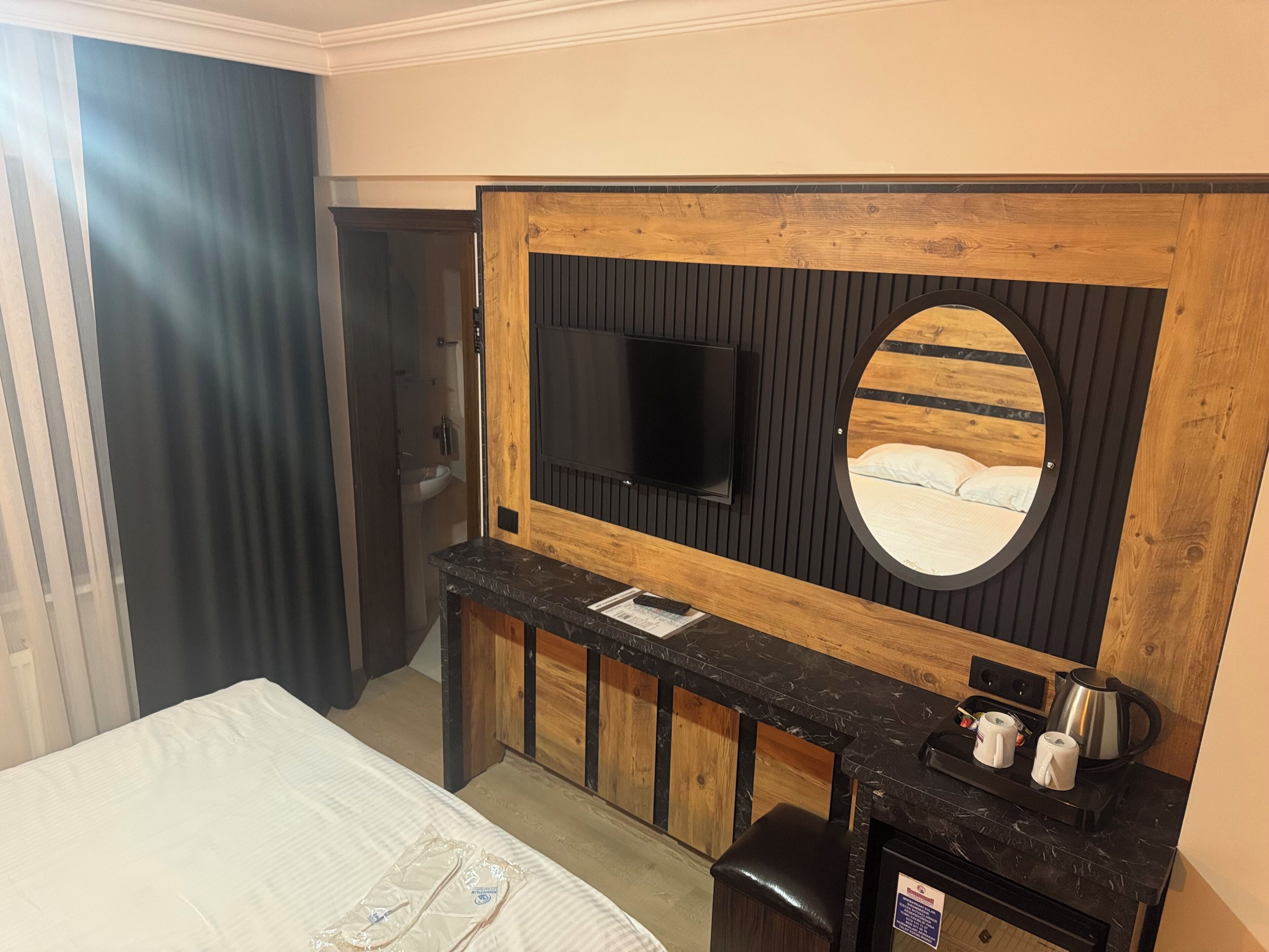 A hotel room with a bed, a desk with a mirror, a television mounted on the wall, and a small kitchenette area.
