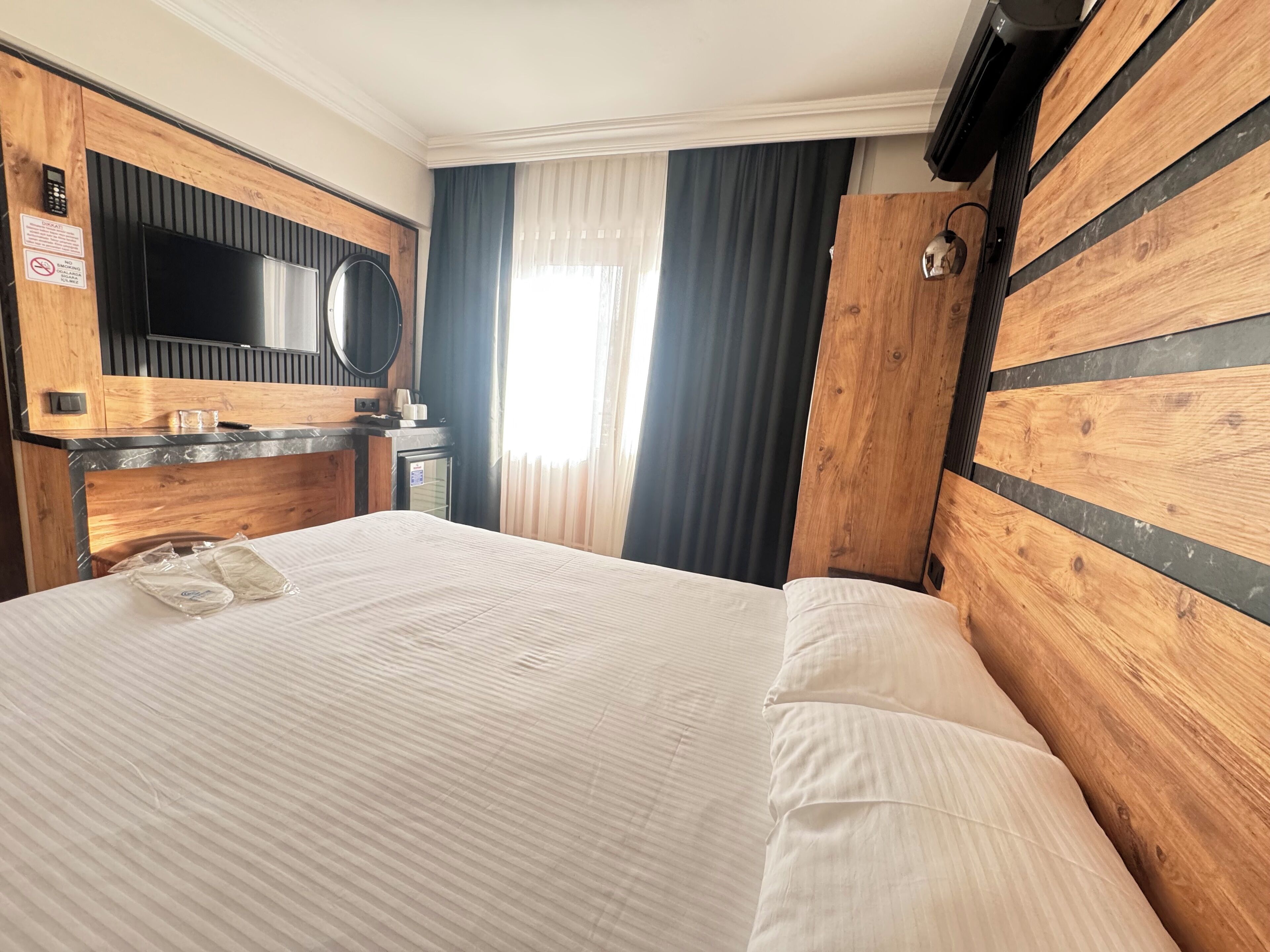 A hotel room with a wooden headboard, a flat-screen TV, a minibar, and a large window with curtains.