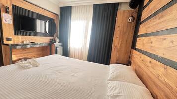 A hotel room with a wooden headboard, a flat-screen TV, a minibar, and a large window with curtains.