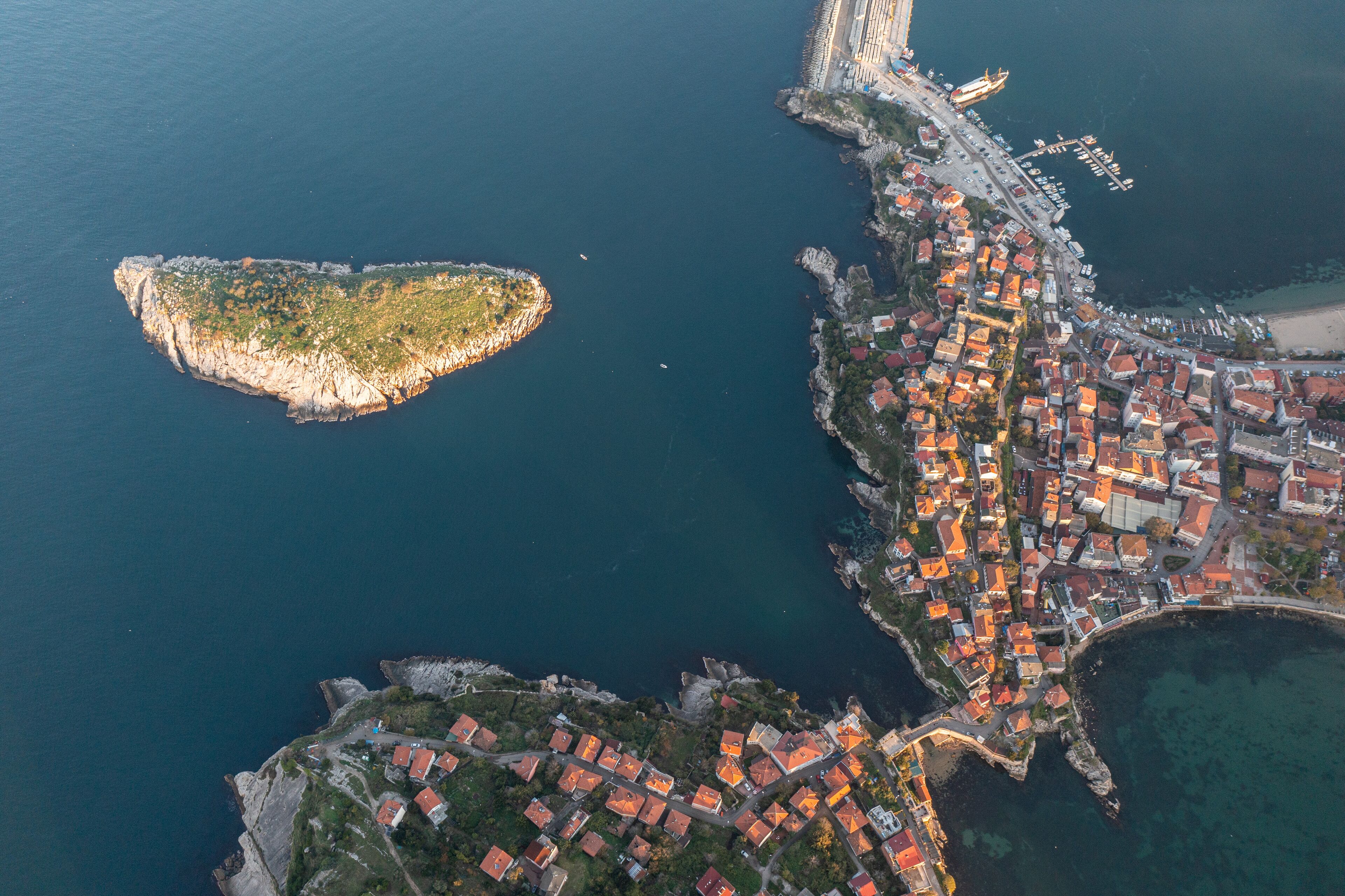 An aerial view of a coastal town with a prominent peninsula, a marina, and a densely packed residential area with red-roofed buildings.
