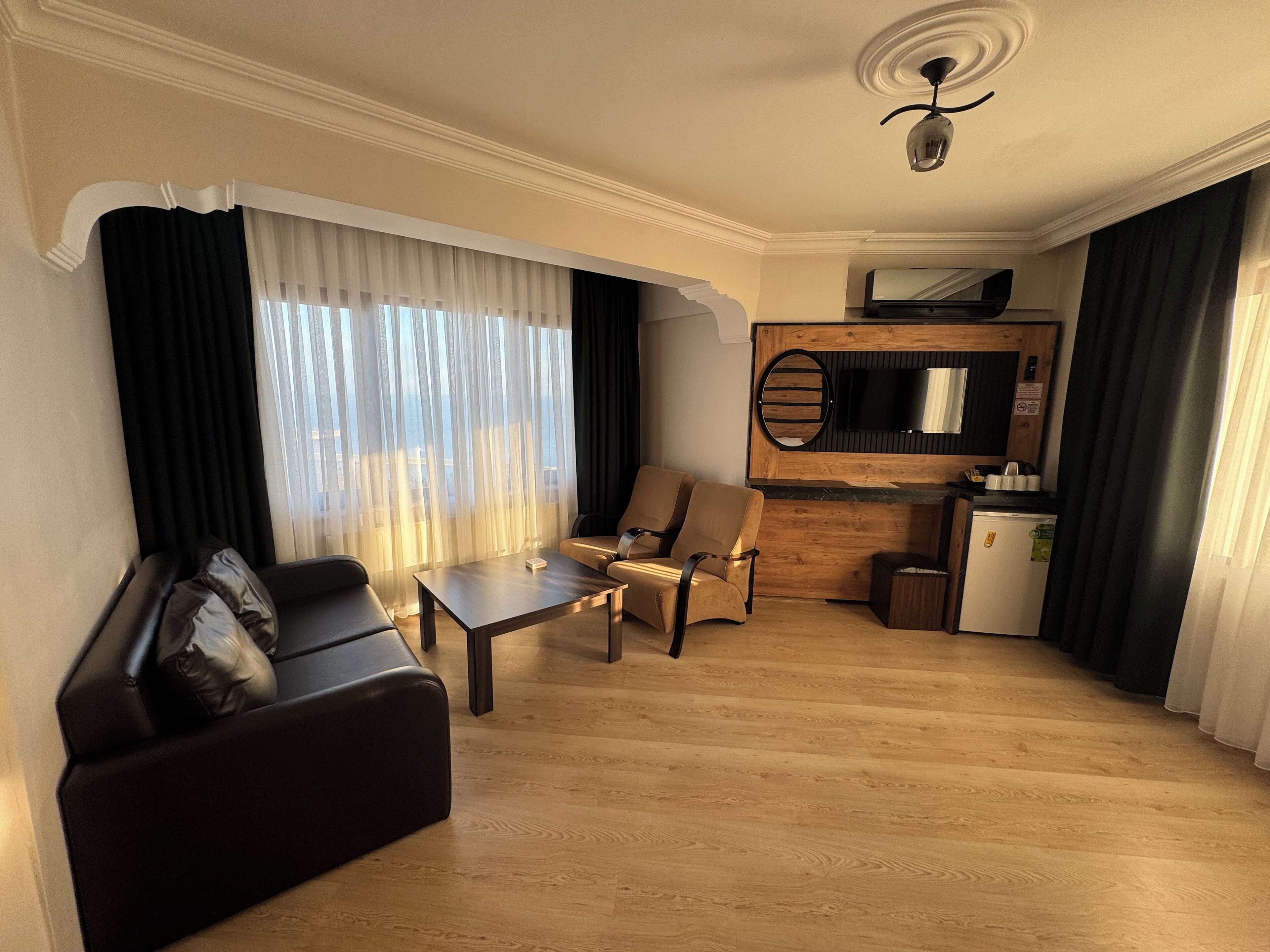 A modern living room with a sofa, a coffee table, and a chair. There is a small kitchenette with a refrigerator and a microwave. The room has large windows with sheer curtains and a view of the city.