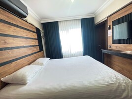 Habitación de hotel con cabecera de madera, una cama grande con ropa blanca, un televisor de pantalla plana en la pared y una ventana con cortinas azules.