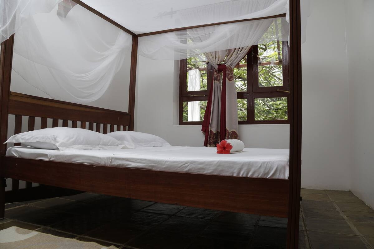 Un lit simple avec un cadre en bois et une literie blanche, une fenêtre avec des rideaux et une vue sur la verdure à l’extérieur.
