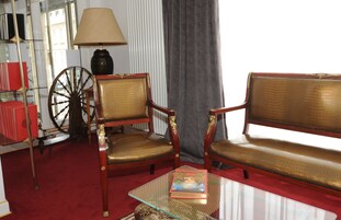 Una habitación con alfombra roja, dos sillas de madera con detalles dorados, una mesa de centro de vidrio, una lámpara, una estantería y un ventanal con cortinas.