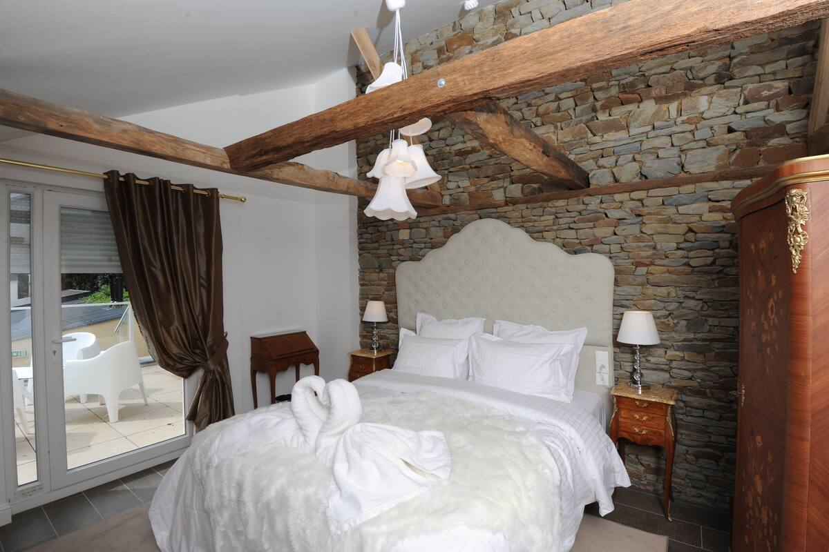 A bedroom with a stone wall, a wooden ceiling, a bed with white bedding, a nightstand with a lamp, and a view of a patio with a table and chairs.