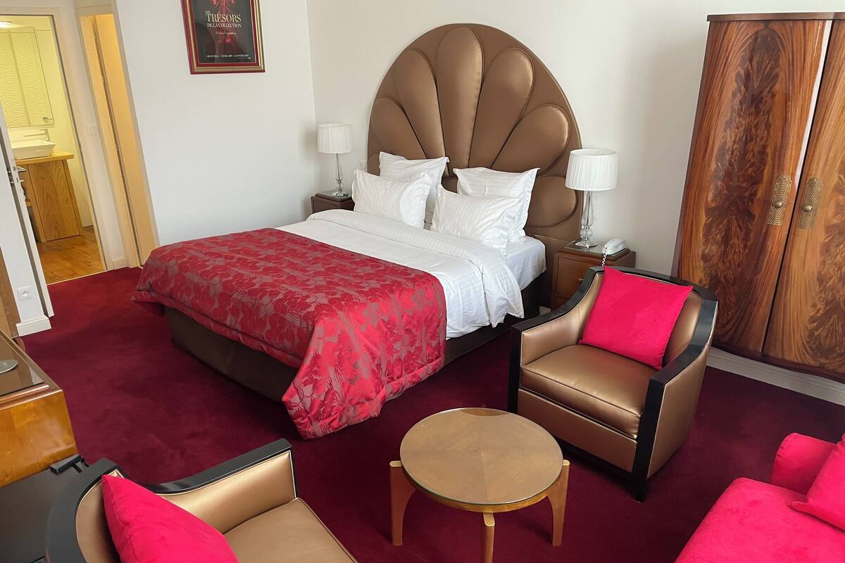 A hotel room with a large bed, two armchairs, a small table, a wooden wardrobe, and a framed picture on the wall.