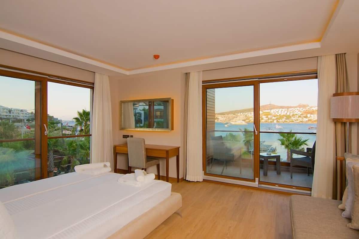 A hotel room with a bed, desk, and large windows offering a view of the sea and cityscape.