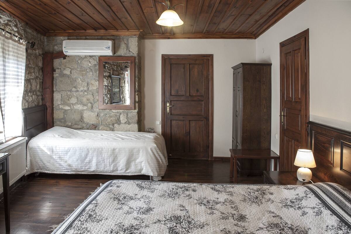 A bedroom with a double bed, a wooden wardrobe, a stone wall, and a window with curtains.