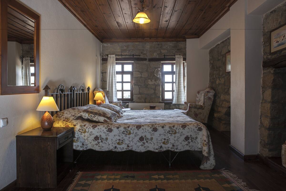 A bedroom with a wooden ceiling, a bed with floral bedding, a nightstand with a lamp, and a stone wall.