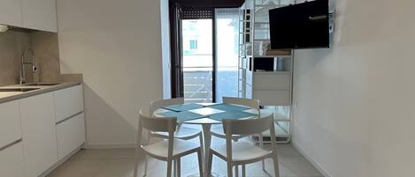 Una cocina compacta con armarios blancos, una mesa de comedor con sillas y un televisor de pantalla plana montado en la pared.