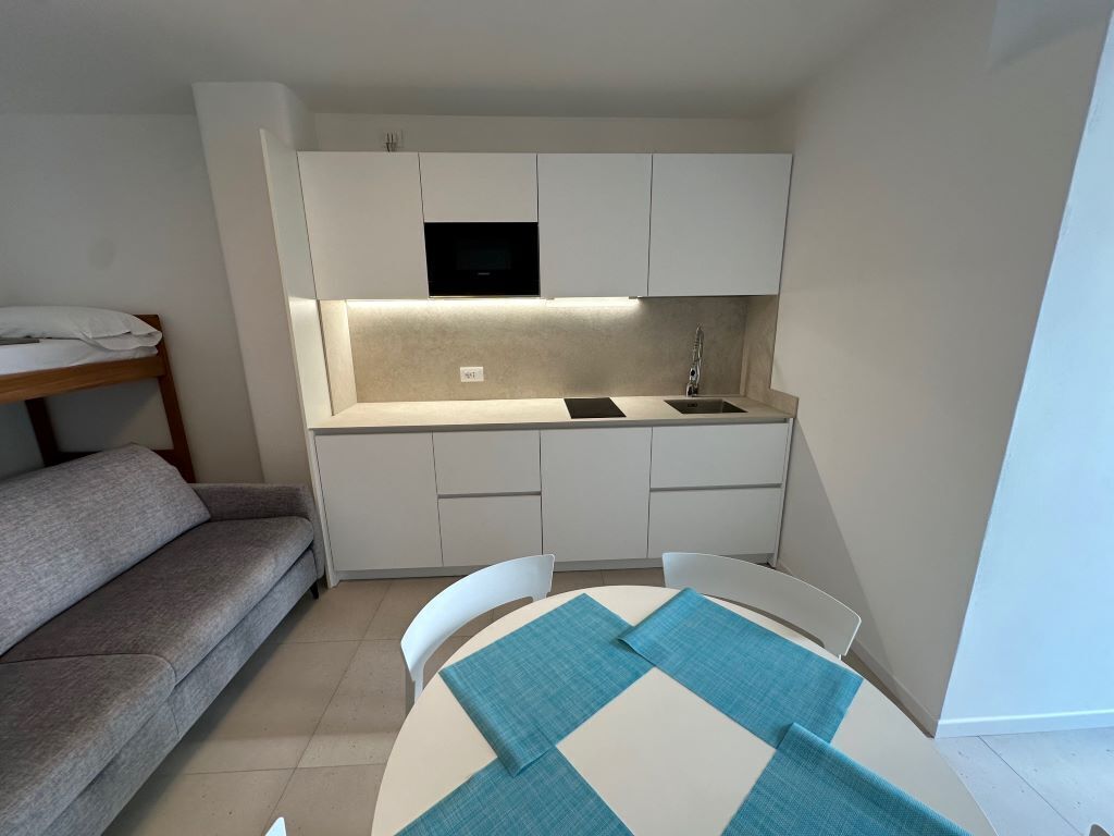 Een compacte keuken met witte kastjes, een ingebouwde magnetron en een spoelbak. Er staat een kleine eettafel met blauwe placemats. Een grijze bank staat links van de keuken.