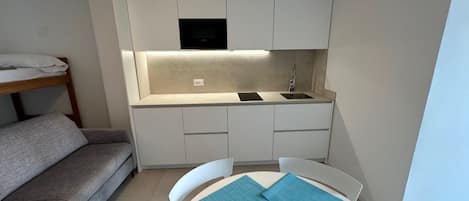 A compact kitchen with white cabinets, a built-in microwave, and a sink. There is a small dining table with blue placemats. A gray sofa is positioned to the left of the kitchen.