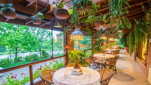 A covered outdoor seating area with wicker tables and chairs, surrounded by hanging plants and potted plants. - Hotel Azaleia (Gramado)