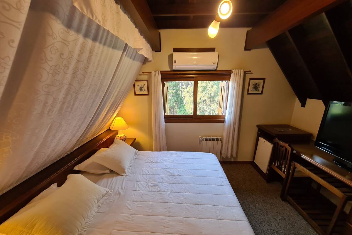 Ein Schlafzimmer mit einem Bett, einer Nachttischlampe, einem Fenster mit Vorhängen, einer Klimaanlage und einem Fernseher auf einem Holzständer.