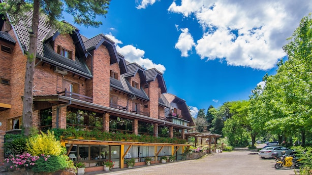 A multi-story brick building with a covered outdoor area, surrounded by greenery and parked cars. - Hotel Azaleia (Gramado)