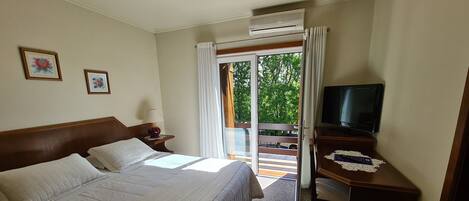 A hotel room with a bed, a television on a wooden stand, and a balcony with a view of trees.