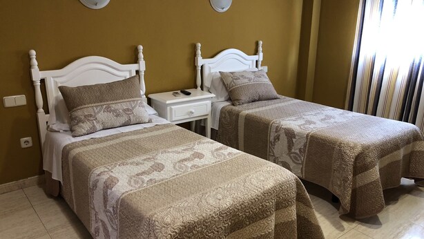 A hotel room with two beds, each with a white headboard and a patterned bedspread. There are two bedside tables with lamps, and a window with vertical blinds.