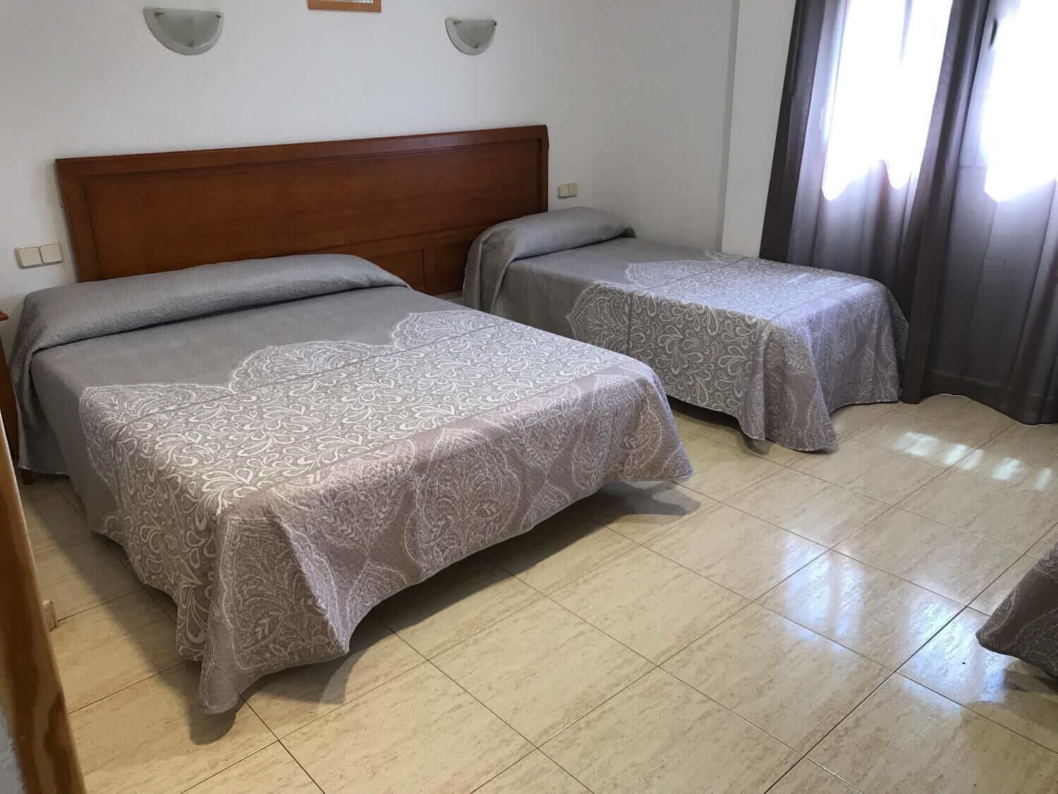 Ein Hotelzimmer mit zwei Betten, einem Holzkopfende, einem Fliesenboden und einem Fenster mit Vorhängen.