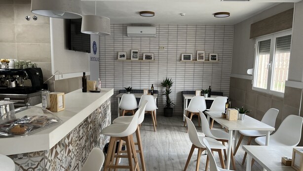 A modern cafe with a counter, white chairs, and a wall-mounted espresso machine. - Hostal 82 (Olias del Rey)