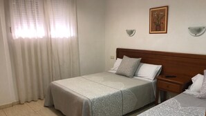 A hotel room with two beds, a wooden headboard, a nightstand, a wall-mounted lamp, and a window with sheer curtains.