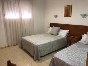 A hotel room with two beds, a wooden headboard, a nightstand, a wall-mounted lamp, and a window with sheer curtains.