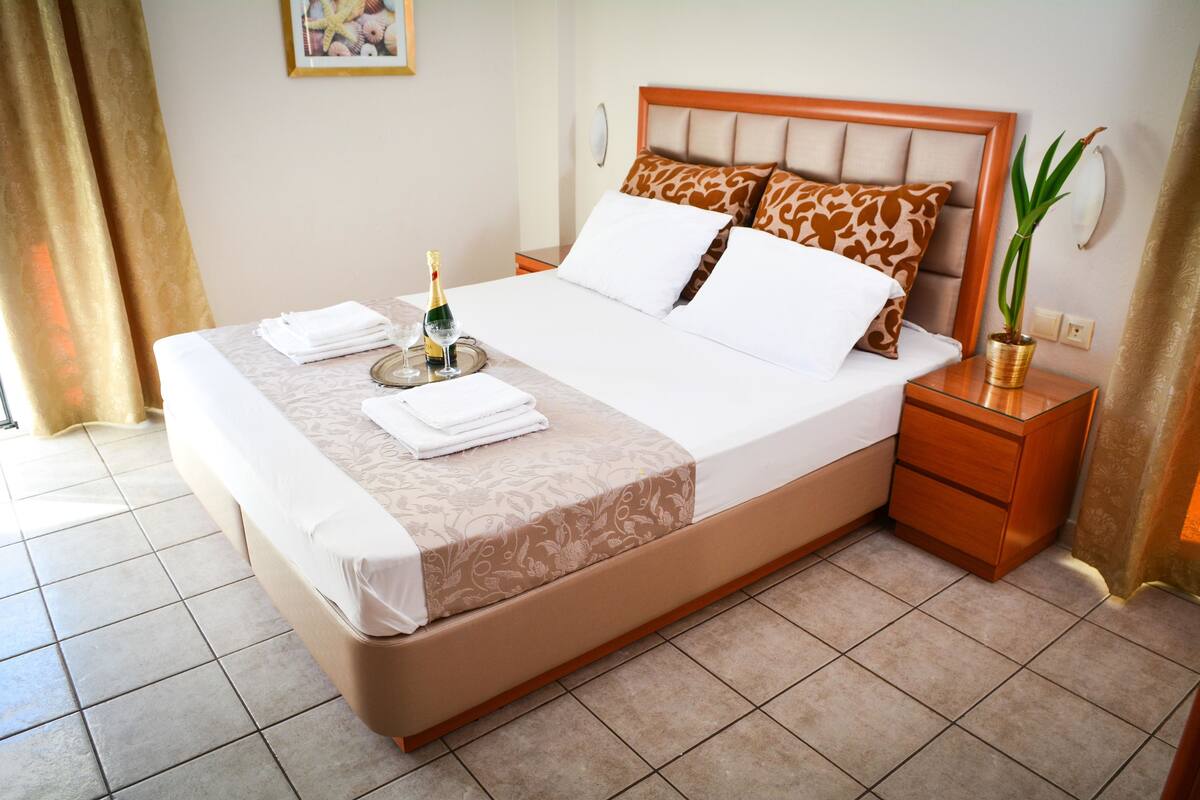 A neatly made bed with a floral bedspread, a bottle of champagne, and folded towels on the side.
