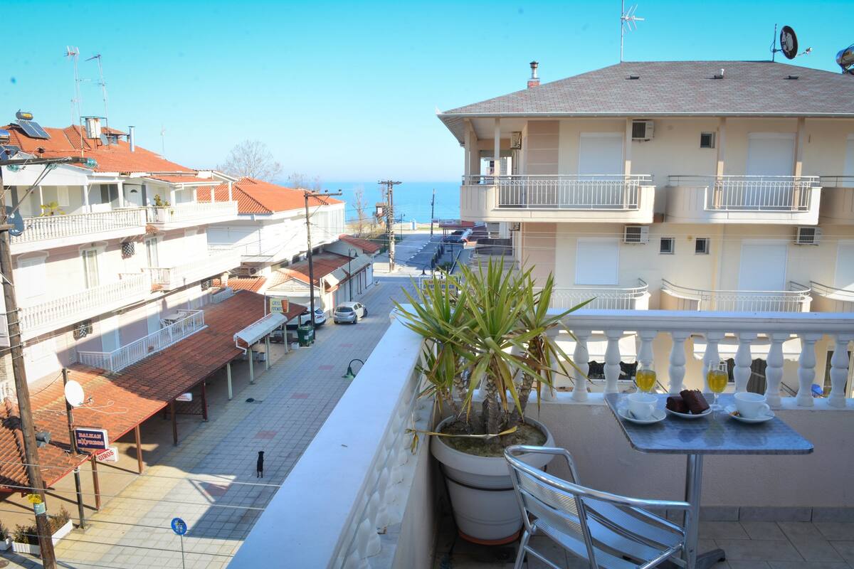 A balcony with a potted plant, a table with a plate of food, and a view of the sea.