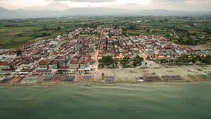 Une vue aérienne d’une ville côtière, avec un ensemble dense de bâtiments, une place centrale ouverte et une zone de plage agrémentée de parasols et de chaises longues.