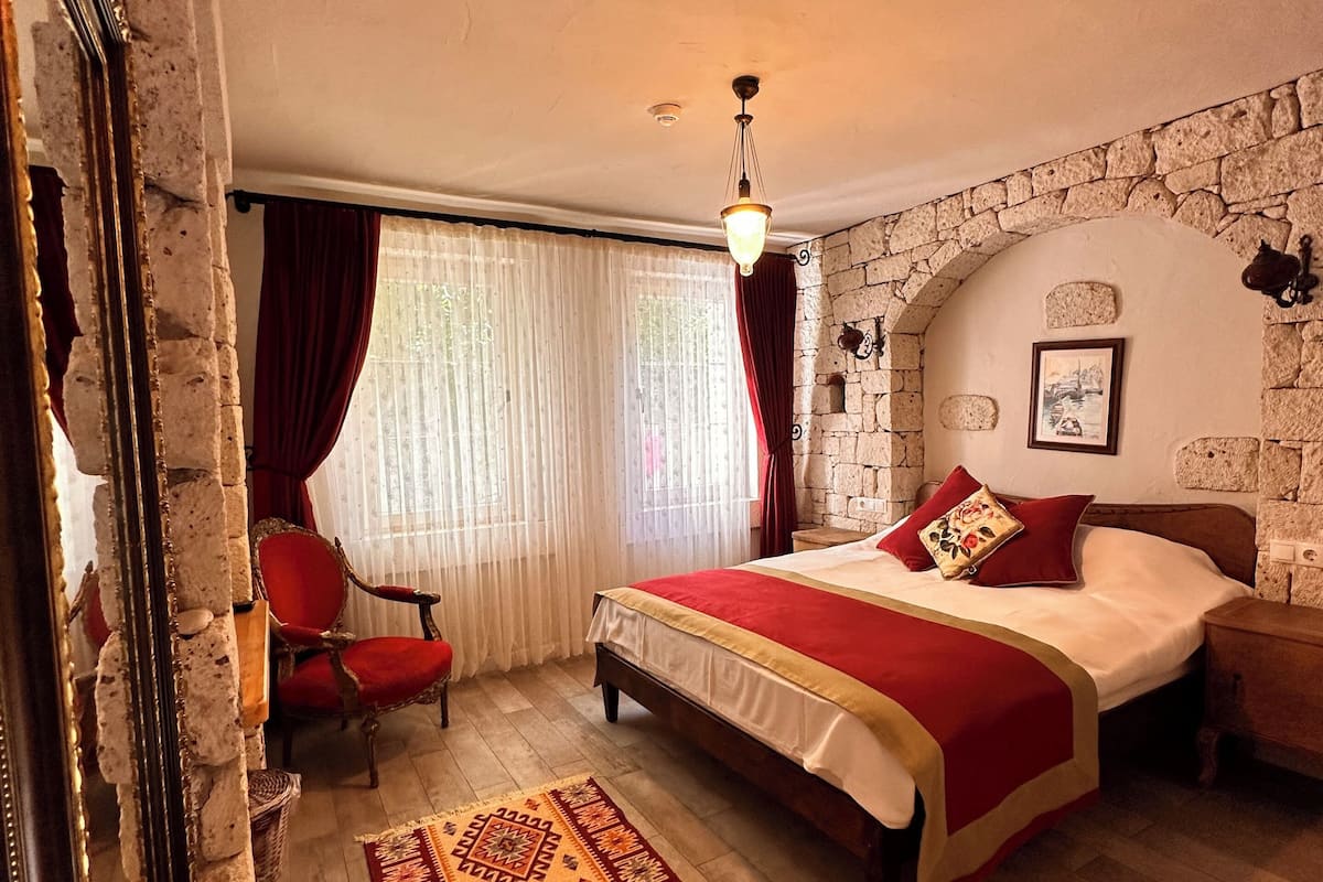 Een slaapkamer met een stenen muur, een bed met rode beddengoed, een rode stoel, een kamerkleed met een patroon en een hanglamp.