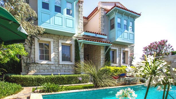 A two-story building with a stone facade and blue window frames, a swimming pool in the foreground, and lush greenery around. - Cassiopeia Hotel Alacati (Cesme)