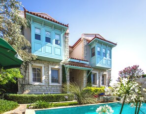 A two-story building with a stone facade and blue window frames, a swimming pool in the foreground, and lush greenery around. - Cassiopeia Hotel Alacati (Cesme)