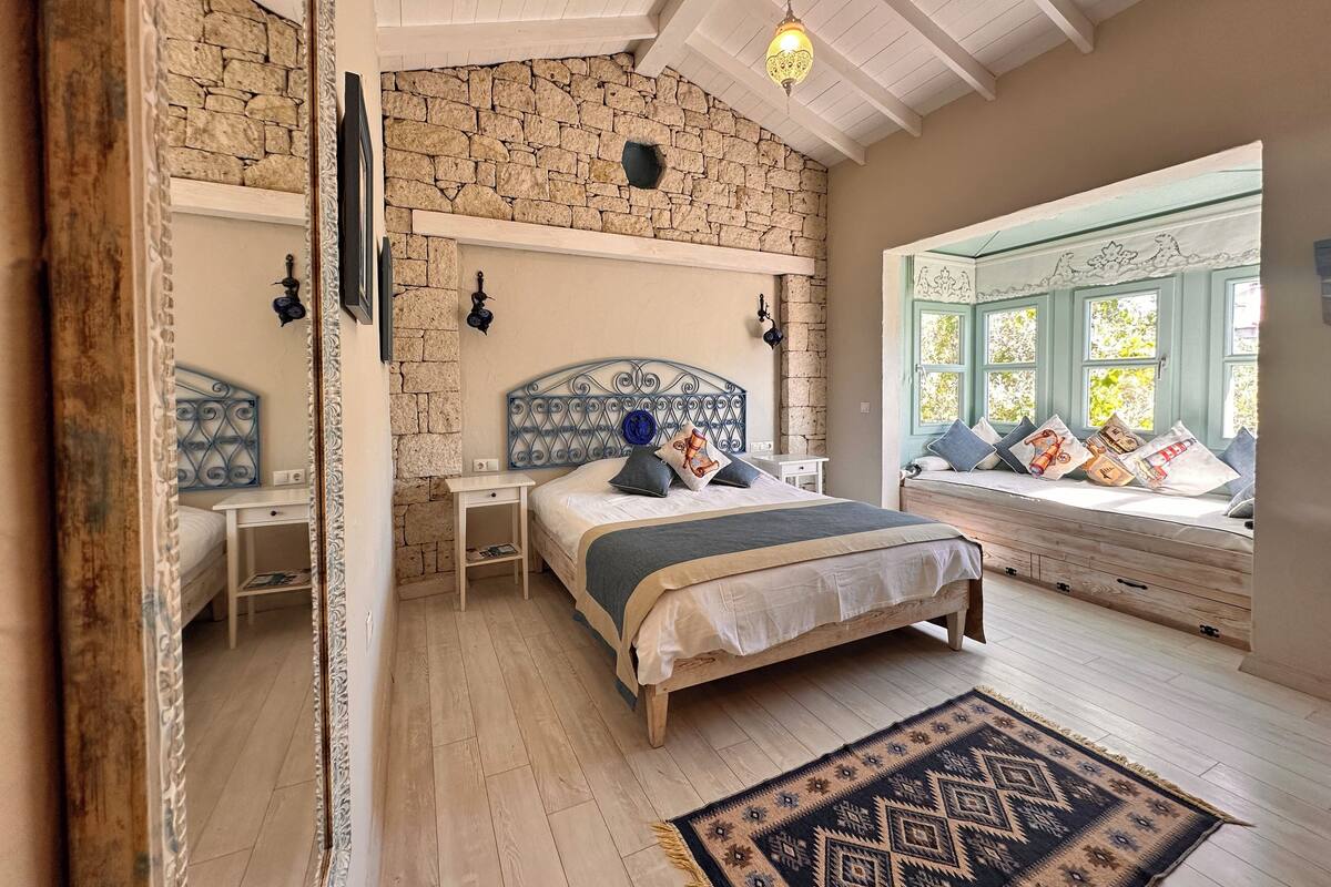 Een slaapkamer met een stenen muur, een bed met een blauw hoofdeinde, een vensterbank met kussens en een kamerbreed tapijt met een patroon.
