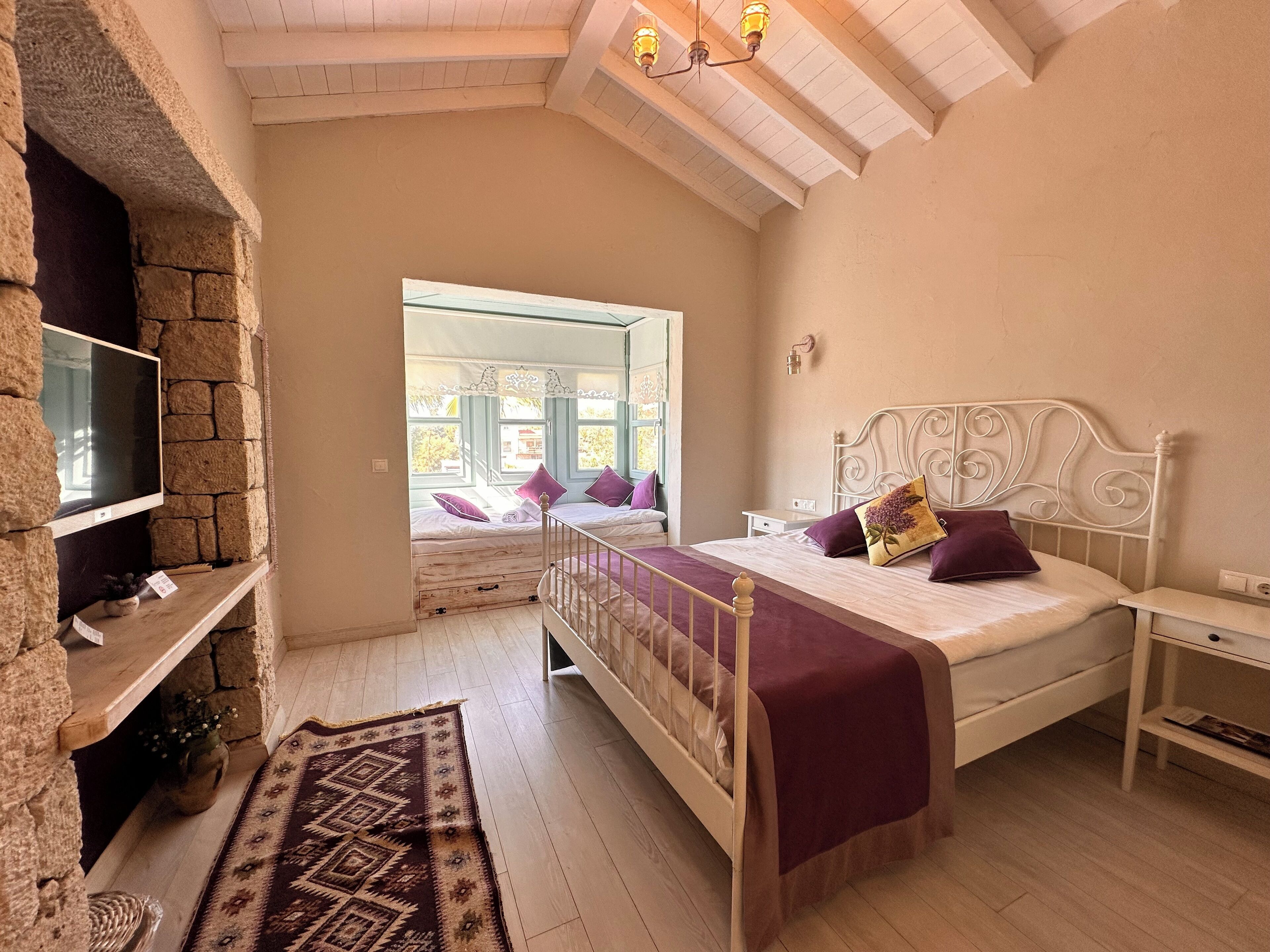 A bedroom with a stone wall, a bed with a headboard, a TV mounted on the wall, a window with a view, and a wooden floor.