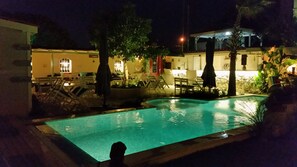 An outdoor swimming pool at night with lounge chairs and tables, surrounded by trees and a building with illuminated windows. - Cassiopeia Hotel Alacati (Cesme)
