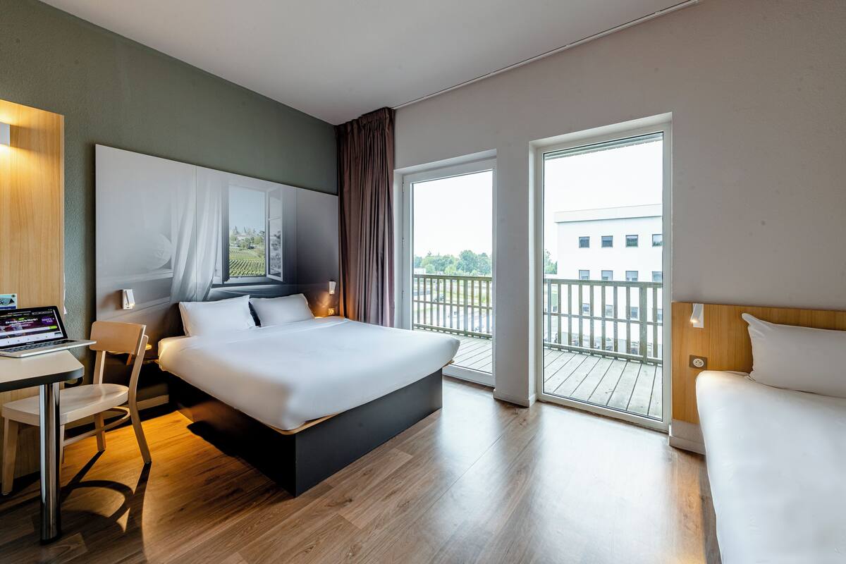 Een moderne hotelkamer met een groot bed, een bureau met een laptop, een stoel en een balkon met uitzicht op de stad.