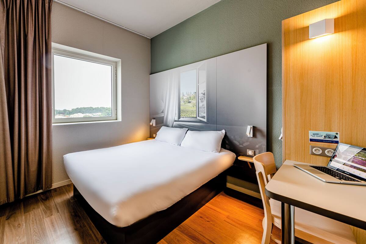 Hotelkamer met een groot bed, een bureau met een laptop, een raam met gordijnen en uitzicht buiten.