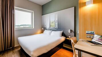 A hotel room with a large bed, a desk with a laptop, a window with curtains, and a view of the outside.
