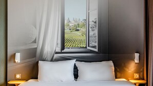 Habitación de hotel con cama, mesita de noche, lámpara de pared y vista a un viñedo por una ventana abierta.