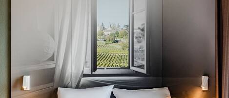 A hotel room with a bed, a nightstand, a wall-mounted lamp, and a view of a vineyard through an open window.