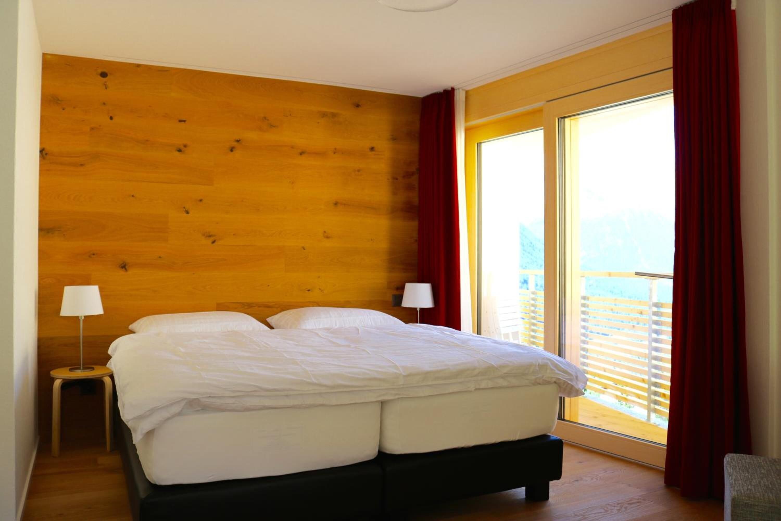 Un dormitorio con pared de madera, una cama con ropa de cama blanca, dos lámparas de noche y un ventanal con cortinas rojas.