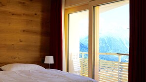 Une chambre à coucher avec une tête de lit en bois, un lit recouvert de linge de lit blanc, une table de chevet avec une lampe et une grande porte-fenêtre coulissante donnant sur un balcon avec vue sur les montagnes.