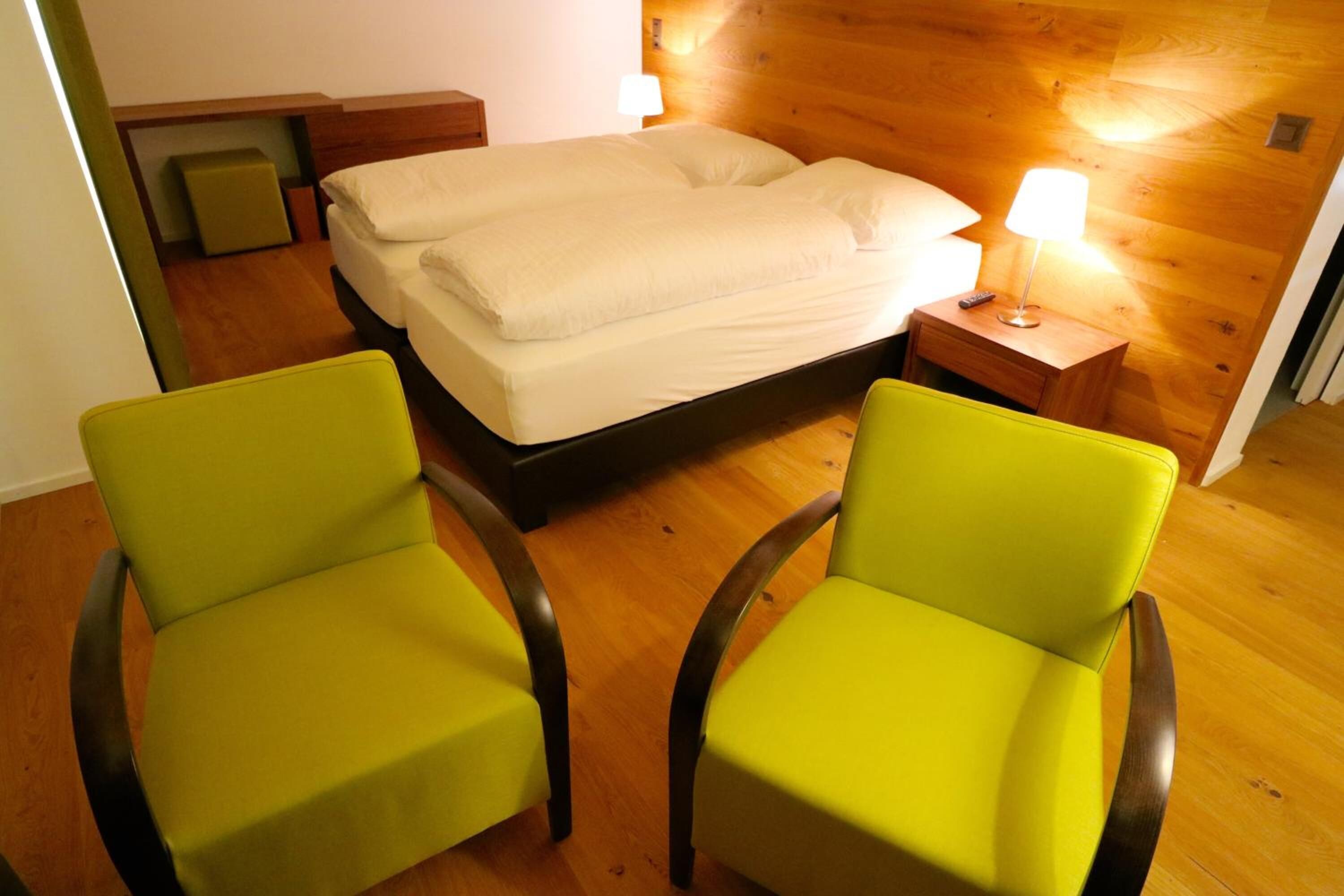 Ein Zimmer mit einem Bett, zwei grünen Sesseln, einem Nachttisch mit Lampe und Holzwänden.