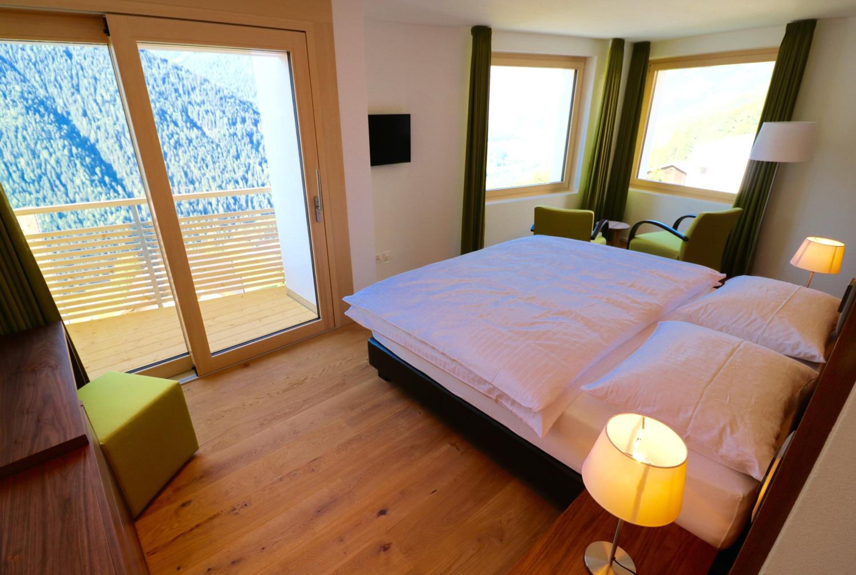 A hotel room with a large bed, a desk, a chair, and a view of a mountainous landscape through the sliding glass door.