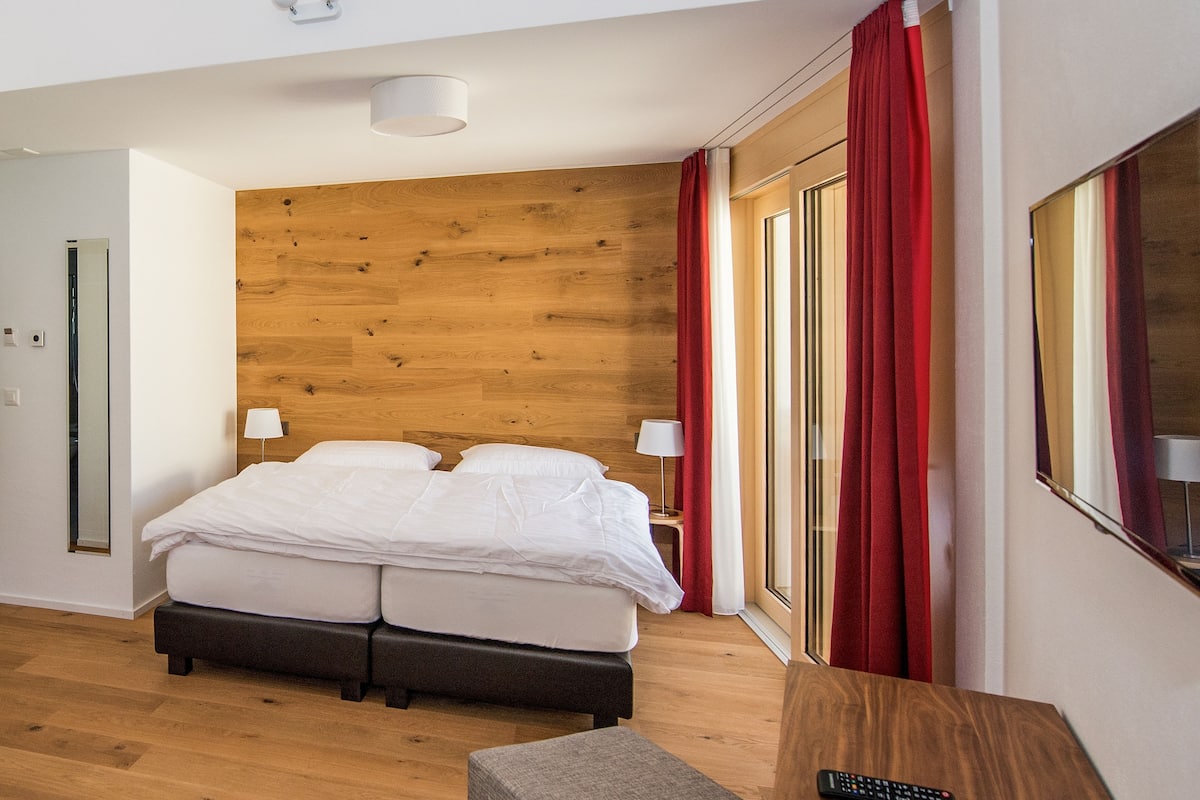 A hotel room with a wooden headboard, a bed with white bedding, a small table with a remote control, and a window with red curtains.