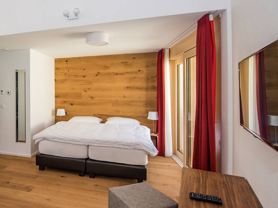 A hotel room with a wooden headboard, a bed with white bedding, a small table with a remote control, and a window with red curtains.