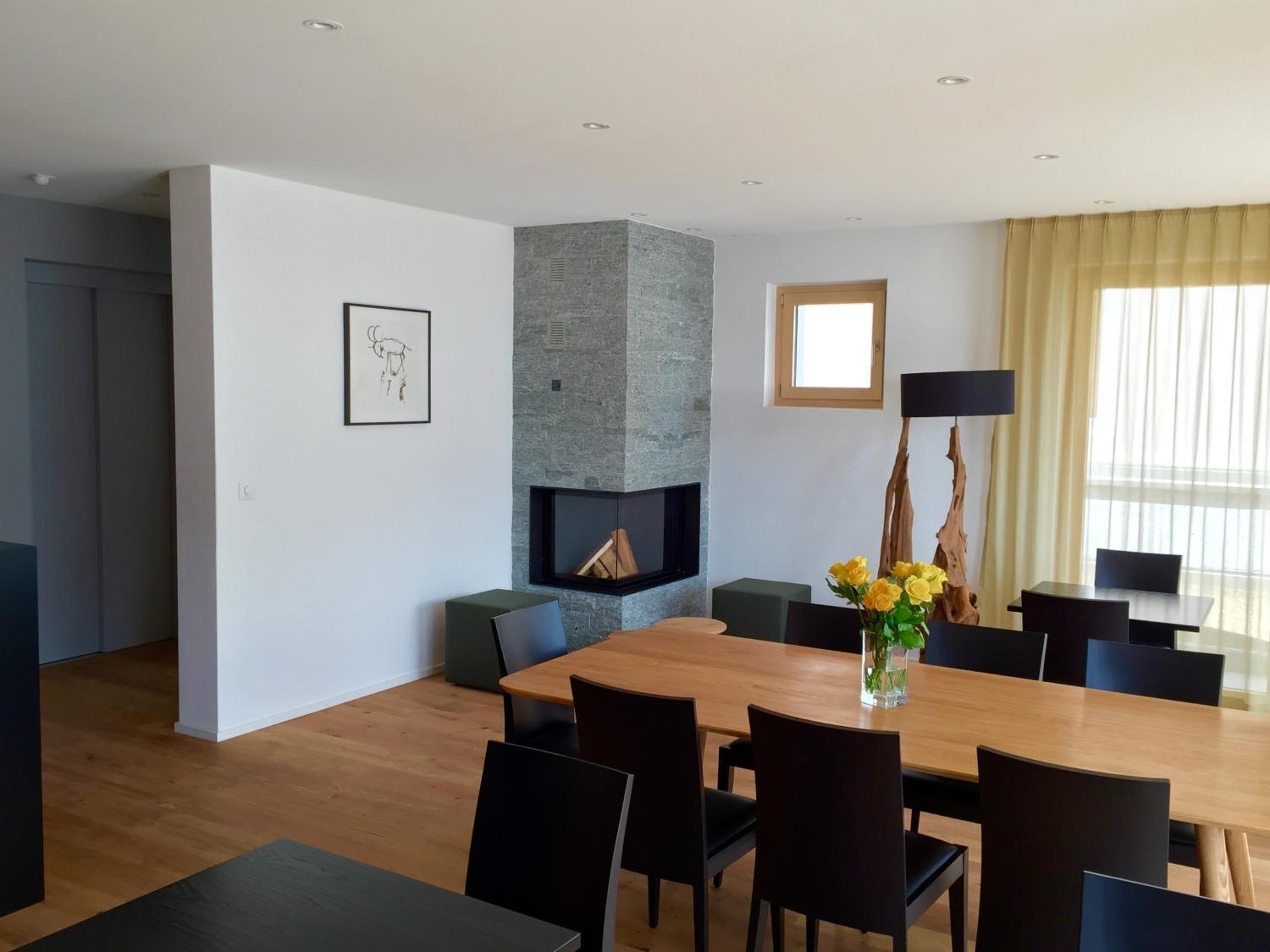 A modern dining area with a wooden table, black chairs, a fireplace, and a large window with curtains.